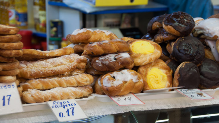 Assorted glazed, sugared, custard-filled, and chocolate pastries arranged on trays at a market counter.