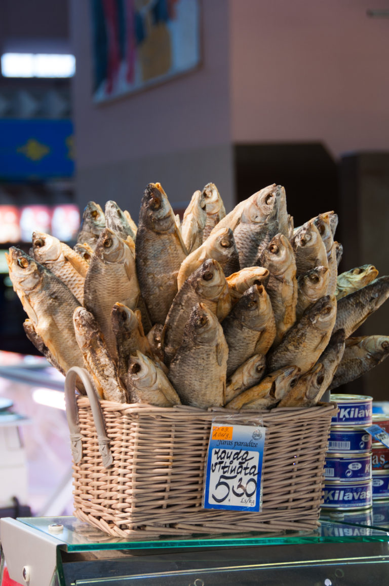 Wicker basket filled with whole dried fish beside a handwritten price card at a Riga market stall.