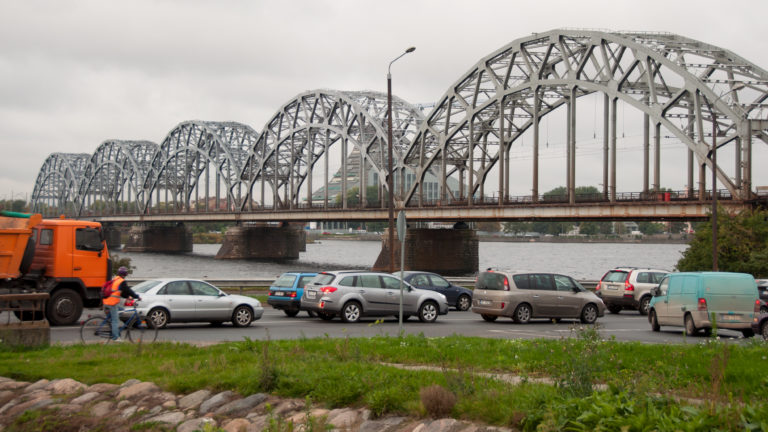 Steel railway bridge across the Daugava behind parked vehicles, streetlights, and a grassy riverside foreground.