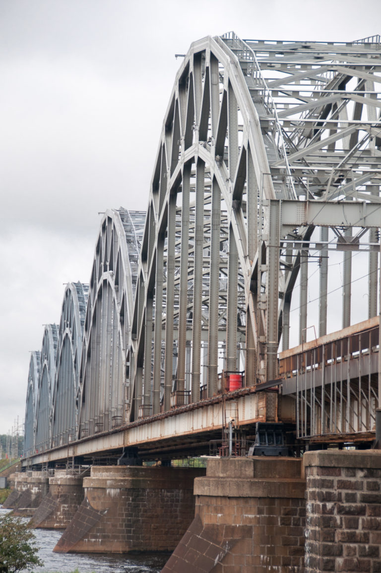 Long steel arch railway bridge crossing the Daugava on masonry piers beneath an overcast sky.