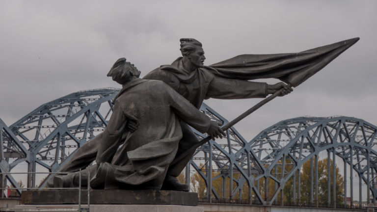 Two monumental figures with a billowing banner stand before the repeating steel arches of Riga’s railway bridge.