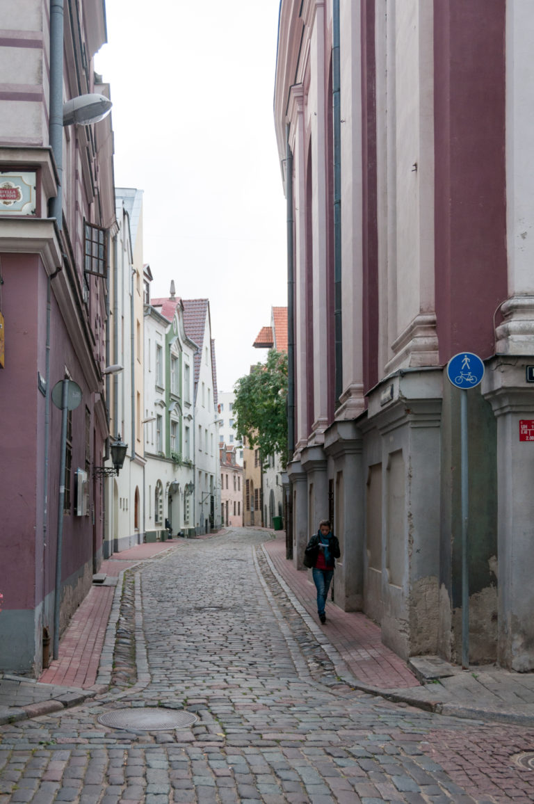 Narrow cobblestone lane in Old Riga between a large church wall and pastel historic buildings, with one pedestrian.