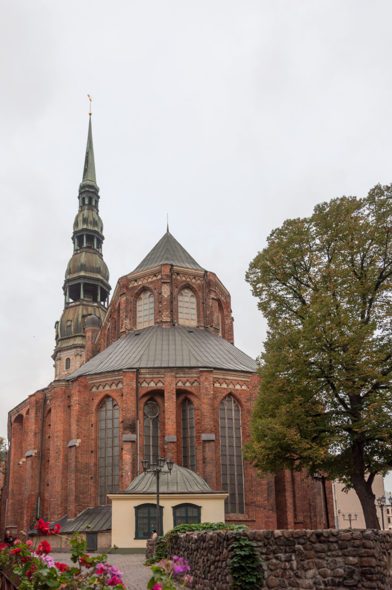 Brick polygonal east end of St. Peter’s Church with tall Gothic windows, landscaped foreground, and copper tower behind.