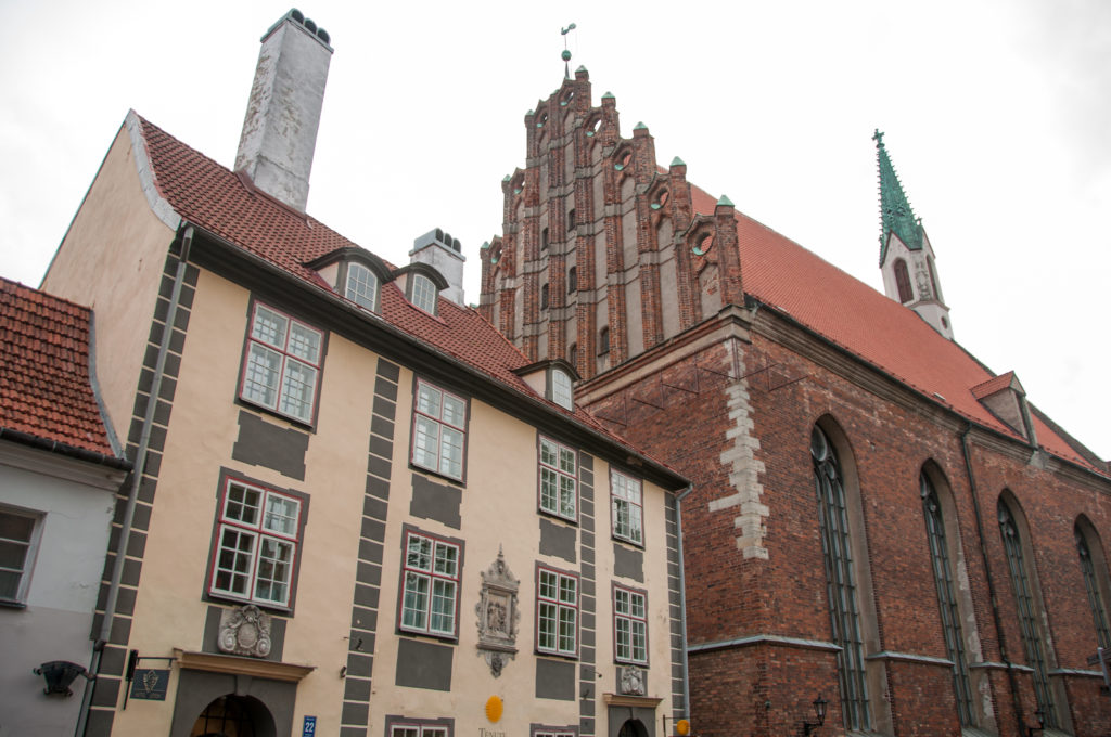 Historic residence with painted trim standing beside the tall red-brick Gothic walls and stepped gable of St. John’s Church.
