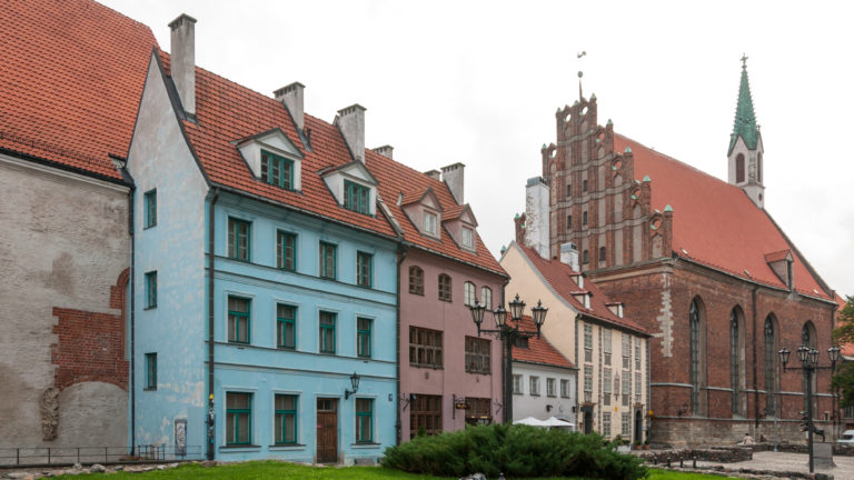 Pastel Old Riga houses beside red-brick St. John’s Church with stepped gable, pointed windows, and steep roof.