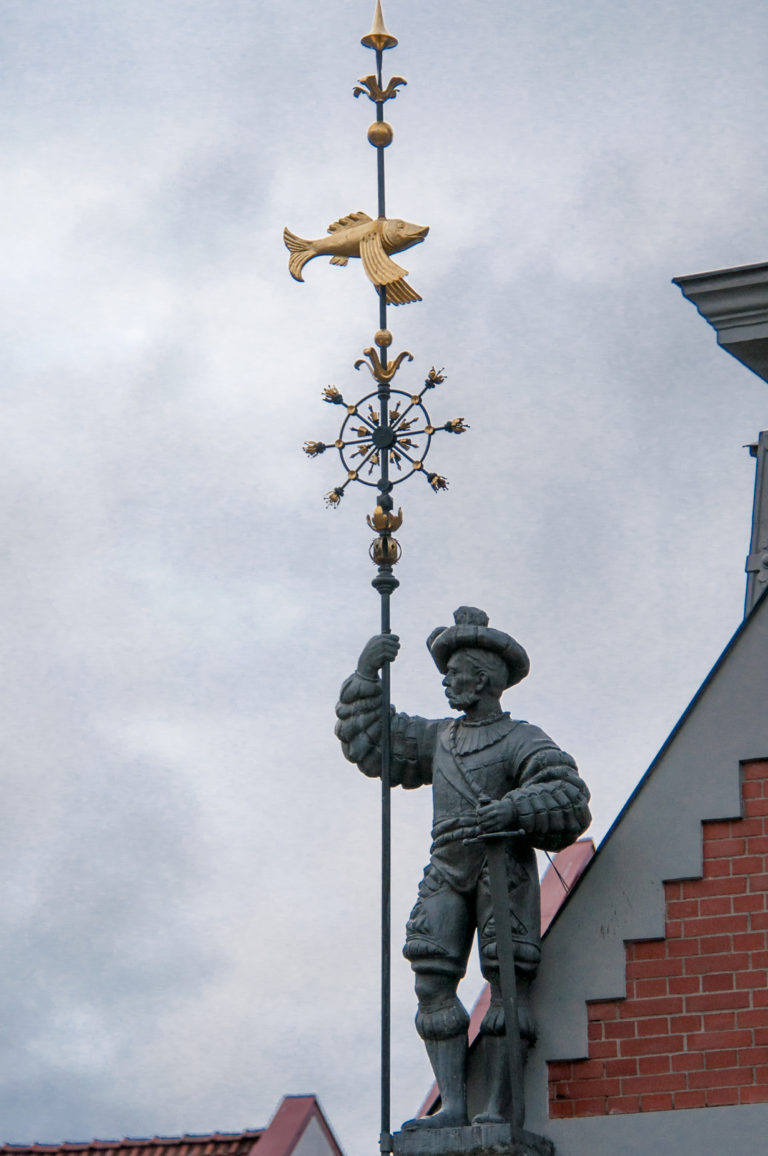 Dark metal guard figure beside a tall finial topped by a gilded bird on a stepped roofline.