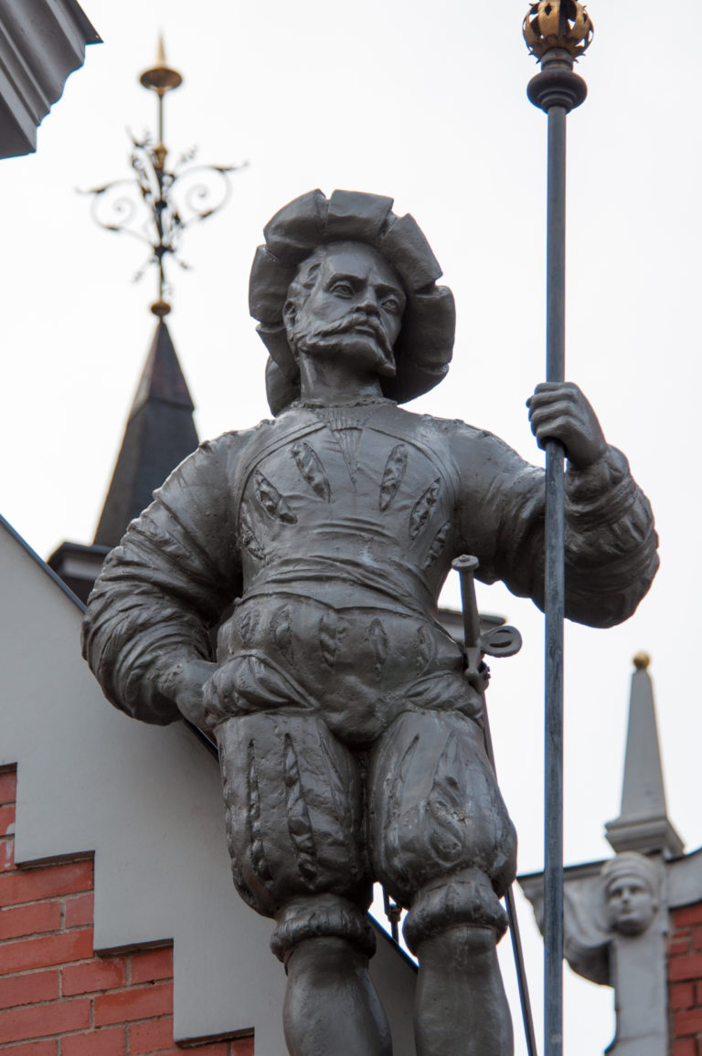 Dark metal Renaissance-style guard figure holding a tall staff above the roofline of the House of the Blackheads.