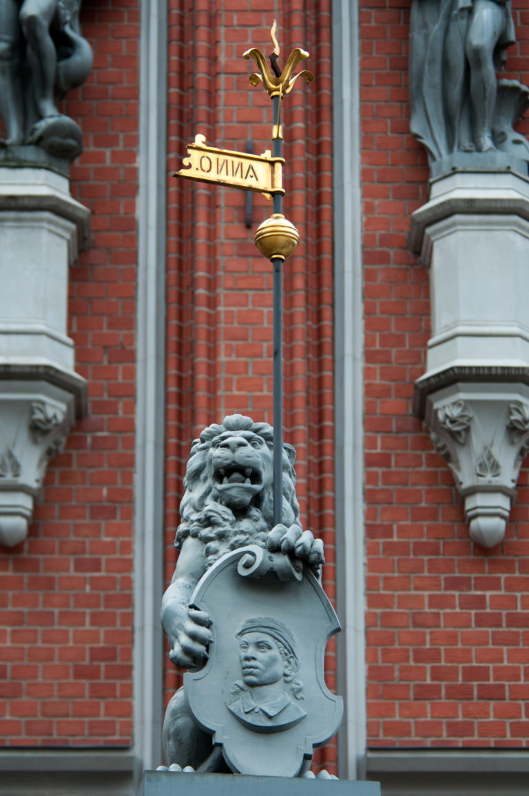 Stone lion holding a circular shield below a gilded banner finial on the House of the Blackheads façade.