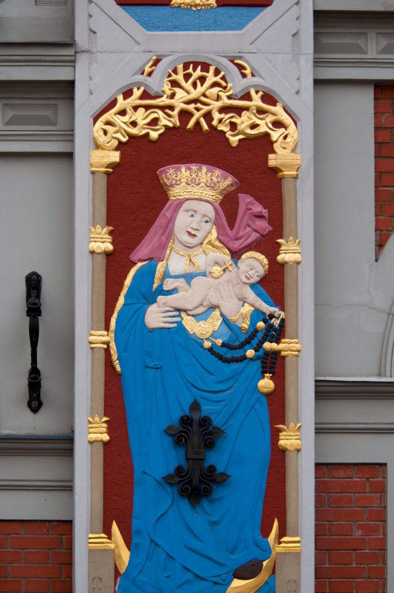 Polychrome crowned Madonna in blue holding the Christ Child within a gilded frame on a red-brick façade.