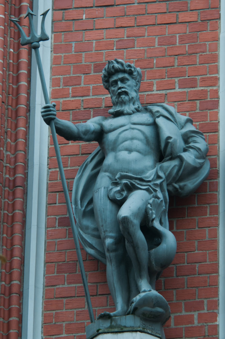 Bearded dark metal male figure holding a long staff on a pedestal against a red-brick façade.