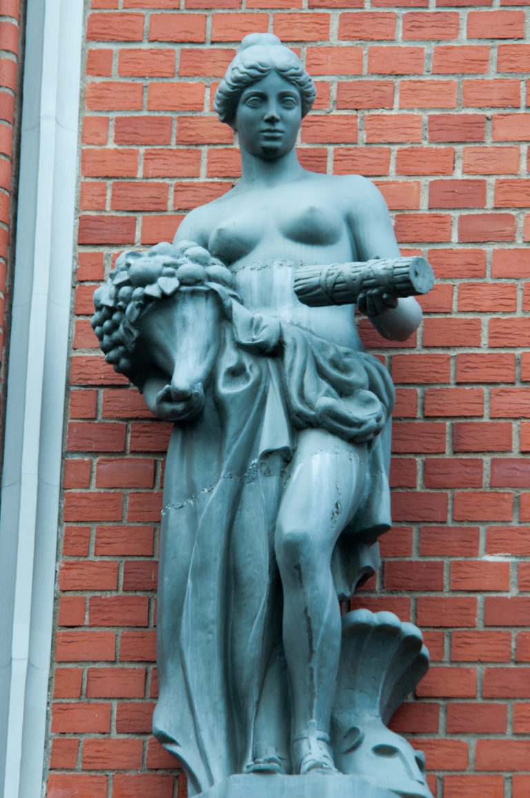 Dark metal female figure carrying fruit or flowers and a cylindrical object against a red-brick façade.