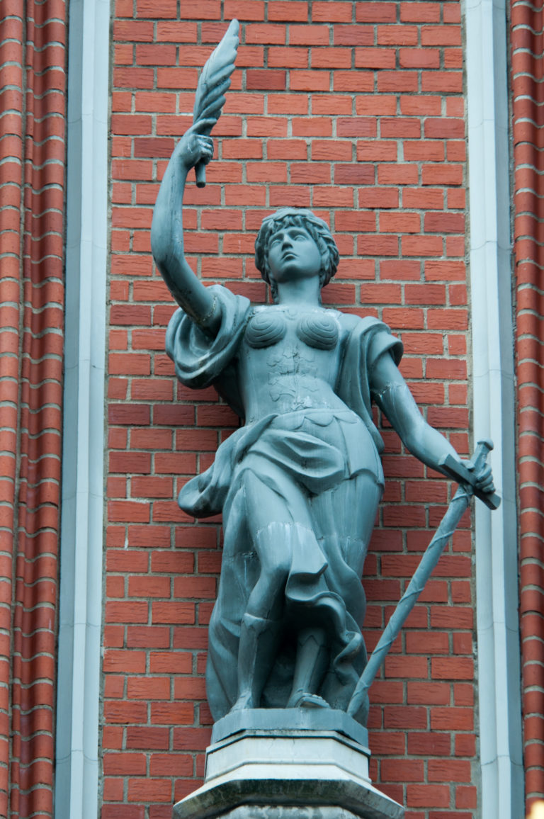 Dark metal female figure with raised arm and long staff mounted on a pedestal against red brick.