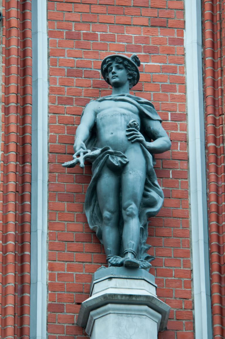 Dark metal classical male figure standing on a pedestal against the red-brick House of the Blackheads façade.