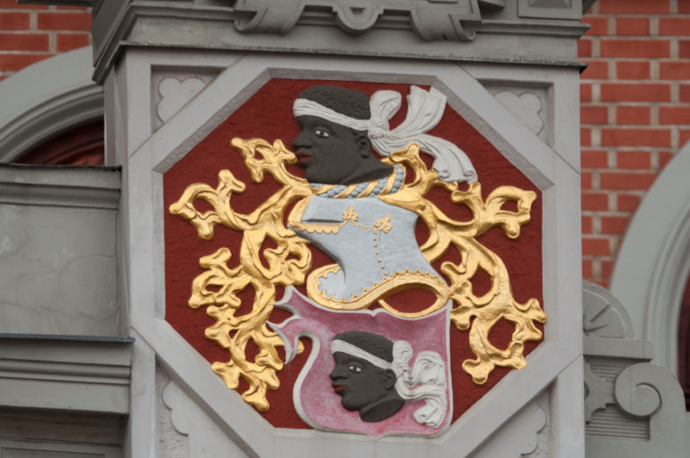 Polychrome coat of arms with profile heads, a turbaned crest, and gold mantling on a red-brick façade.