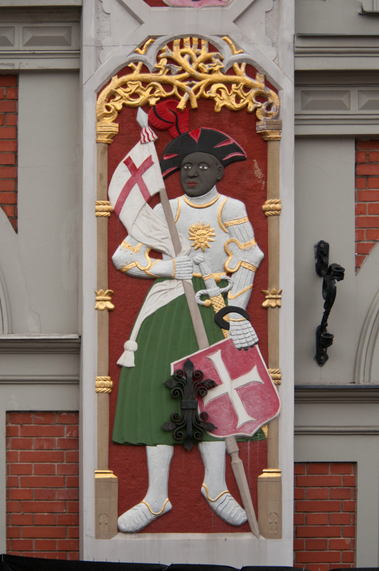 Polychrome armed figure holding a sword and red-and-white shield within a gilded frame on a historic façade.