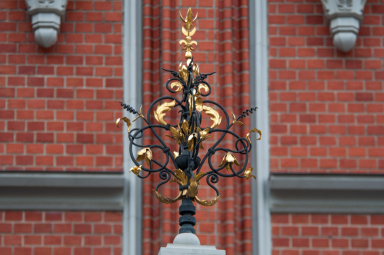 Black wrought-metal finial with gilded leaves and floral details set against a red-brick wall.