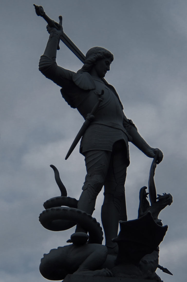 Low-angle silhouette of Saint George with raised sword standing over a dragon against a cloudy sky.