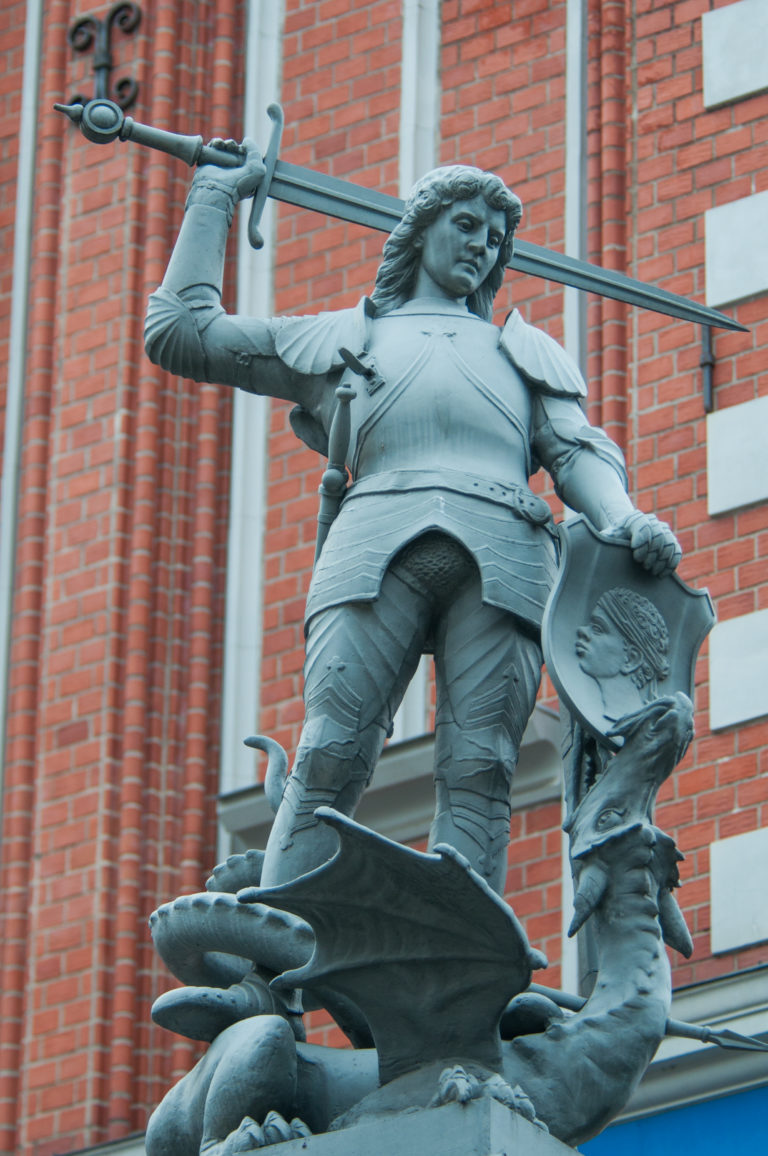 Dark metal sculpture of armored Saint George raising a sword over a twisting dragon on a red-brick façade.