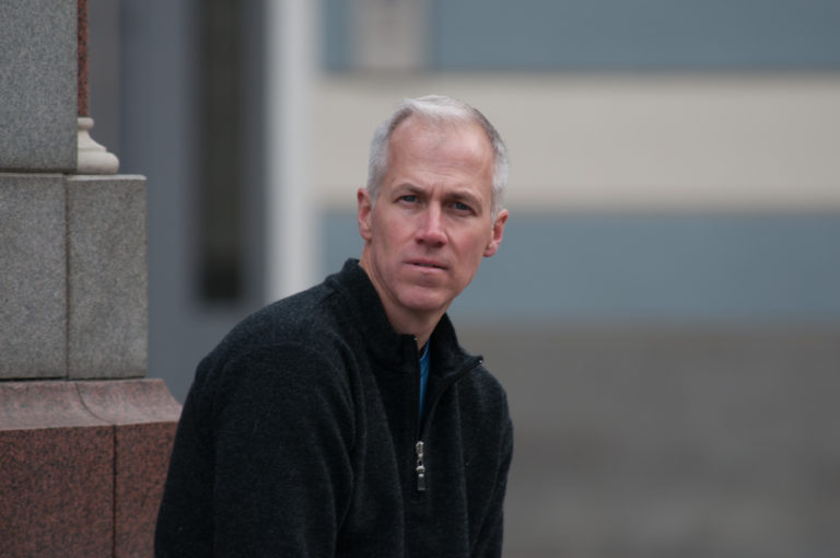 Man in a dark fleece standing beside a stone building corner with a blurred Riga streetscape behind.