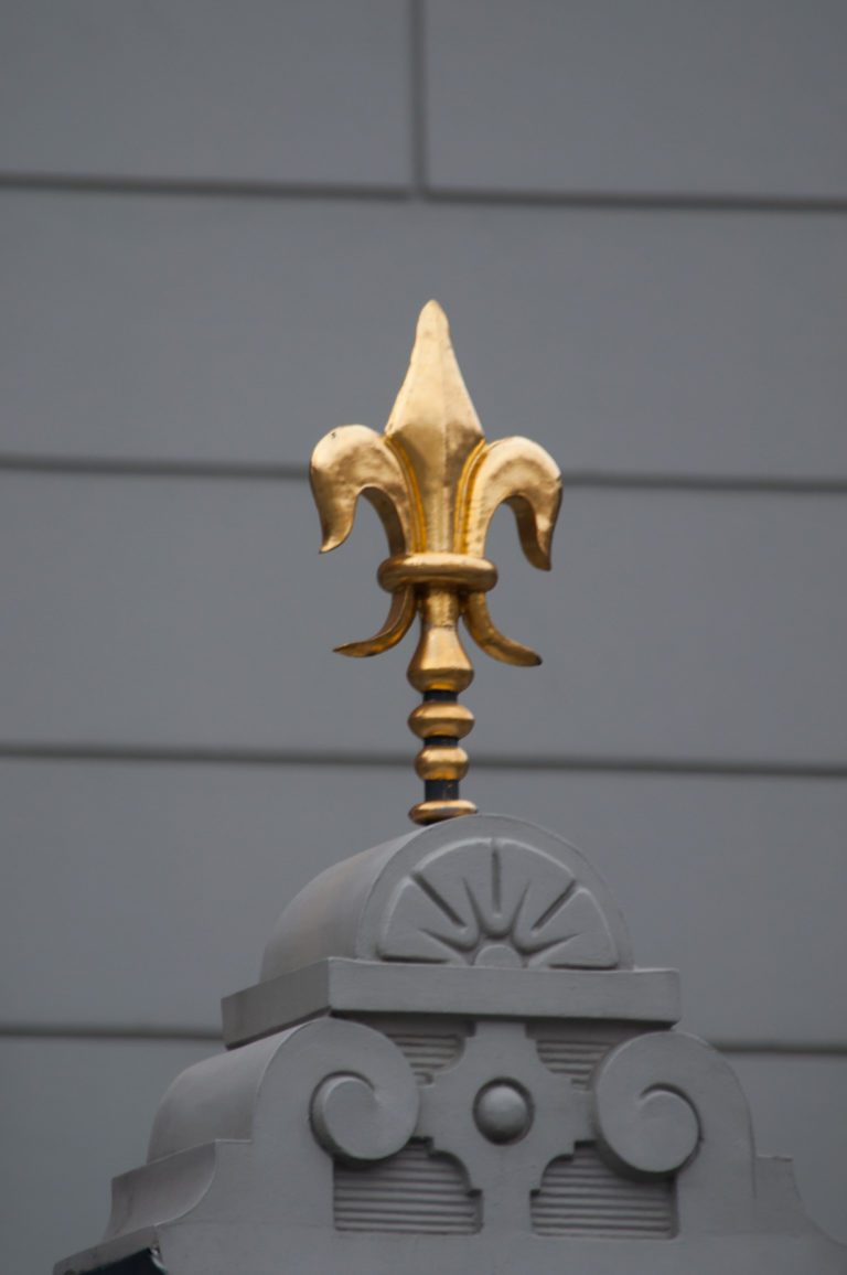 Gilded fleur-de-lis finial mounted above a small decorative pediment on a gray facade.