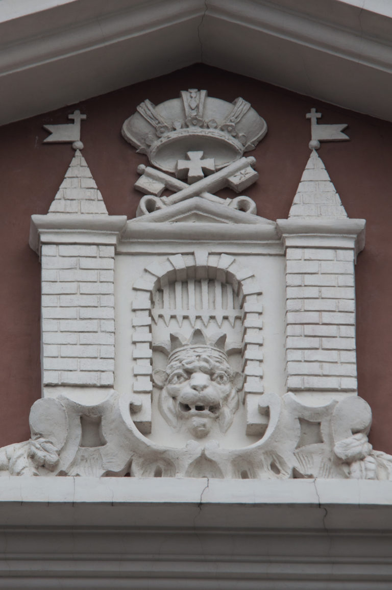 White stucco relief with crowned mask, crossed standards, and stylized towers on a dark red facade.