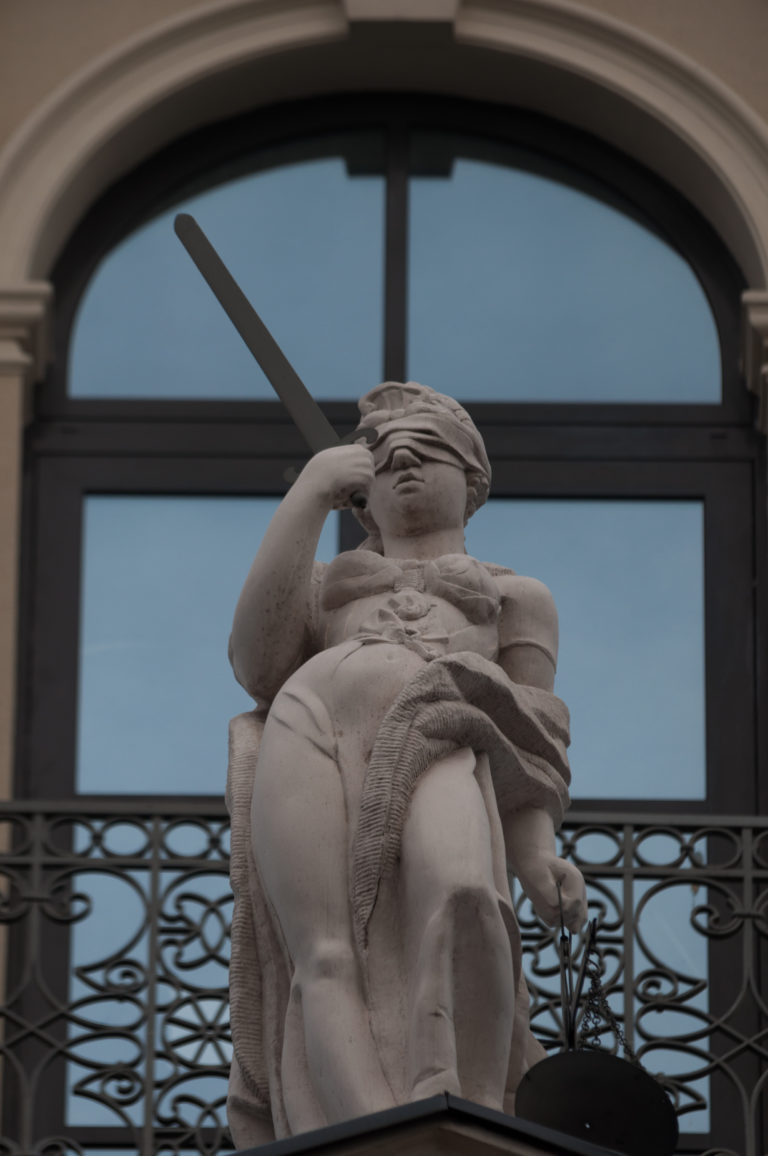 Pale stone sculptural figure set against a dark window and decorative wrought-metal balcony railing.