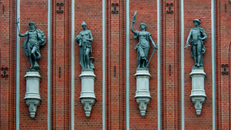 Four standing sculpted figures on brackets against a red-brick historic facade in Old Riga.