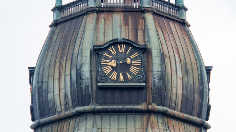 Black clock with gold Roman numerals mounted on a weathered copper-clad tower dome.