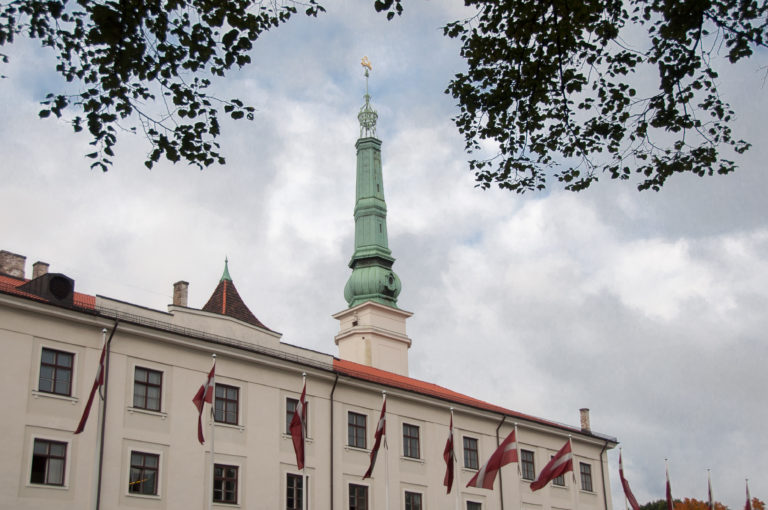 Riga Castle facade lined with Latvian flags and topped by a tall green-copper tower and spire.