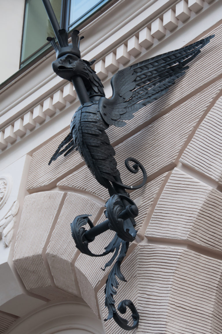 Black metal dragon with spread wings projecting from a pale rusticated building facade.
