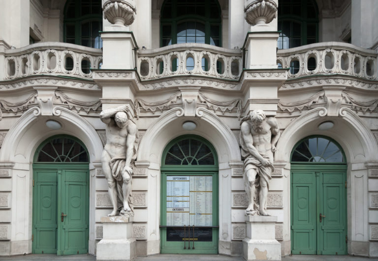 Ornate pale facade with green arched doors, balustrades, and sculpted male atlantes between entrances.