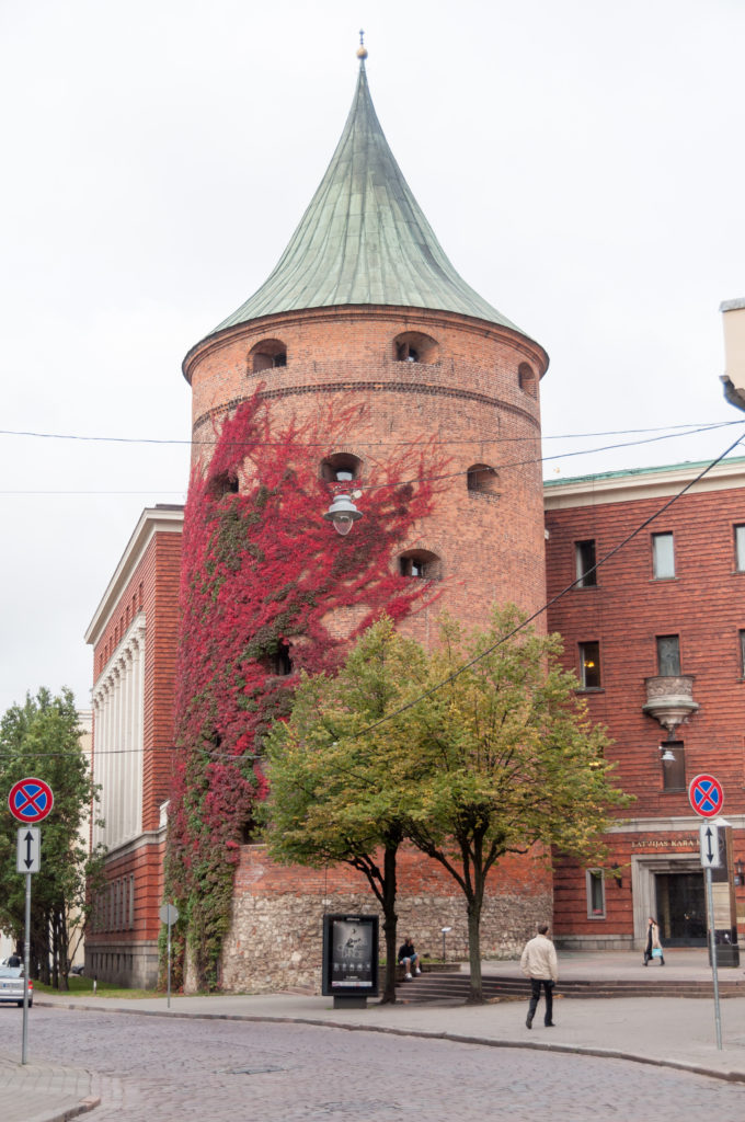 Round brick Powder Tower in Riga with a conical roof, small openings, and red climbing foliage.
