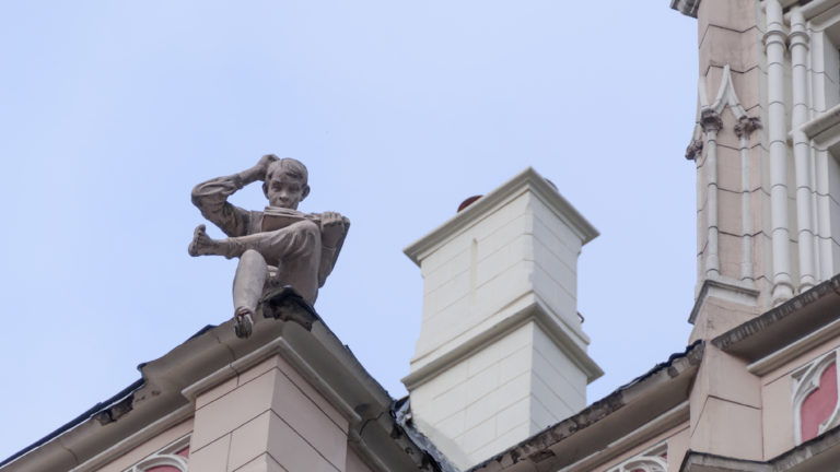 Small rooftop sculpture of a seated violinist beside a tall chimney against the sky.