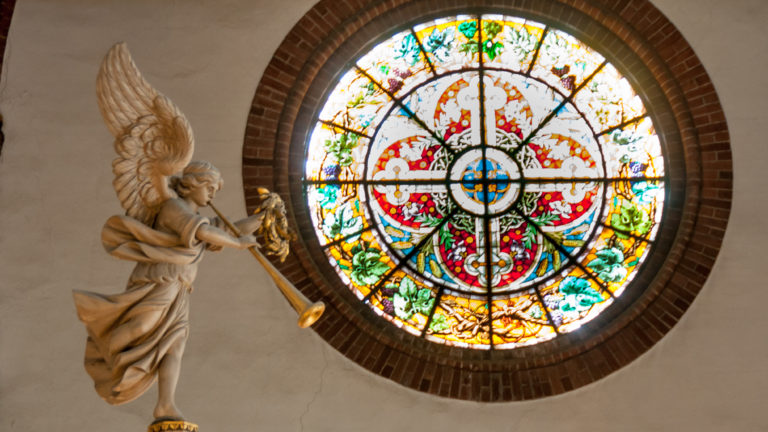 Circular stained-glass window with radial ornament beside a gilded projecting angel sculpture.