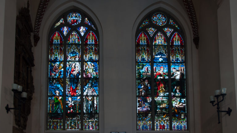 Pair of pointed-arch stained-glass windows with figurative scenes set in a pale church wall.