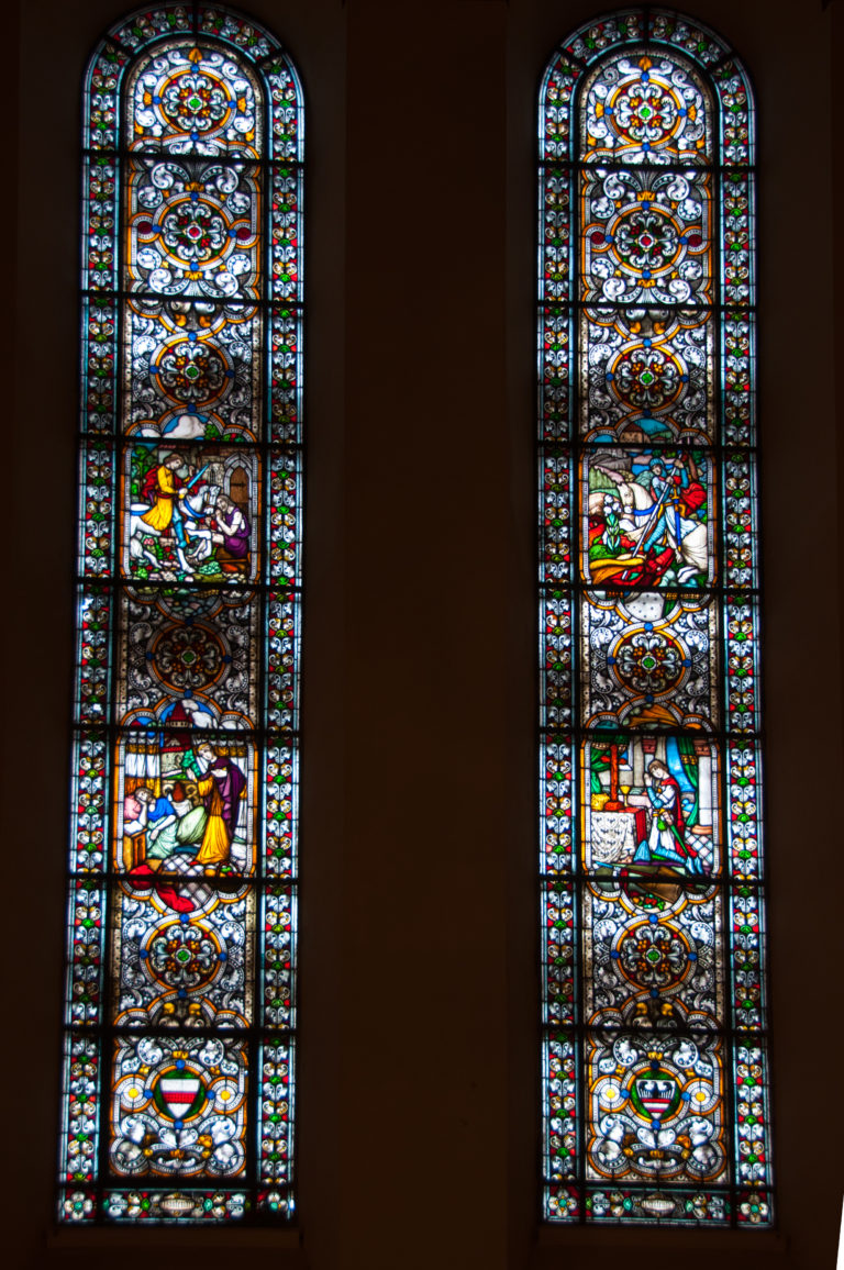 Two tall stained-glass windows with stacked figurative scenes and ornate borders in a dark church interior.