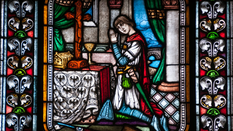 Stained-glass panel with a kneeling figure in prayer beside an architectural setting.