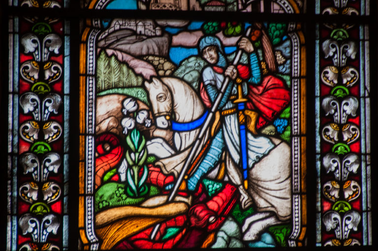 Stained-glass panel with an armored rider on a white horse surrounded by colorful floral borders.