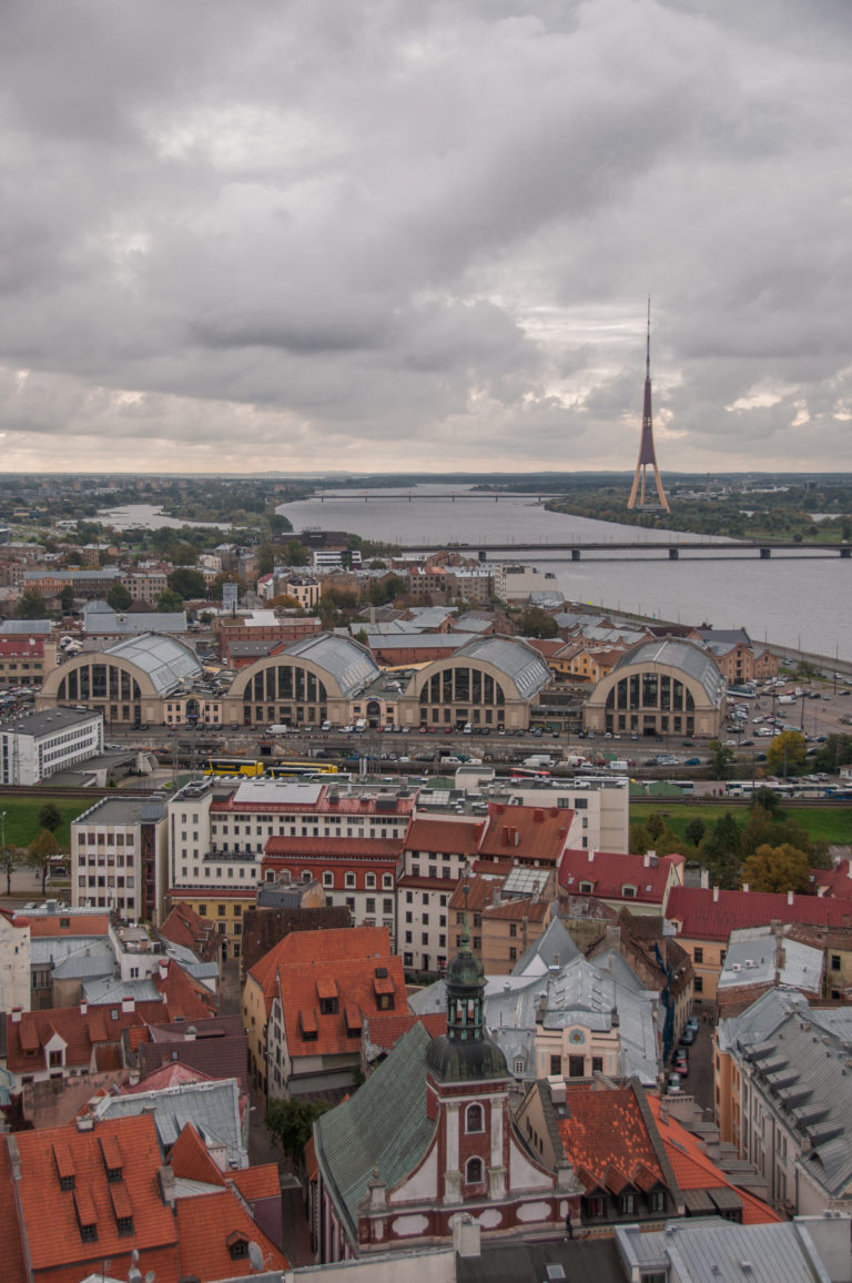 Riga Central Market’s arched halls seen from above, with the Daugava River and Vanšu Bridge beyond.