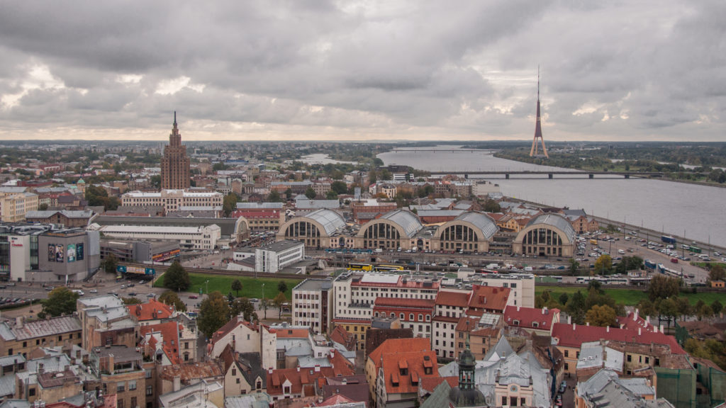 Elevated Riga panorama with Central Market pavilions, Daugava River, and Vanšu Bridge in the distance.
