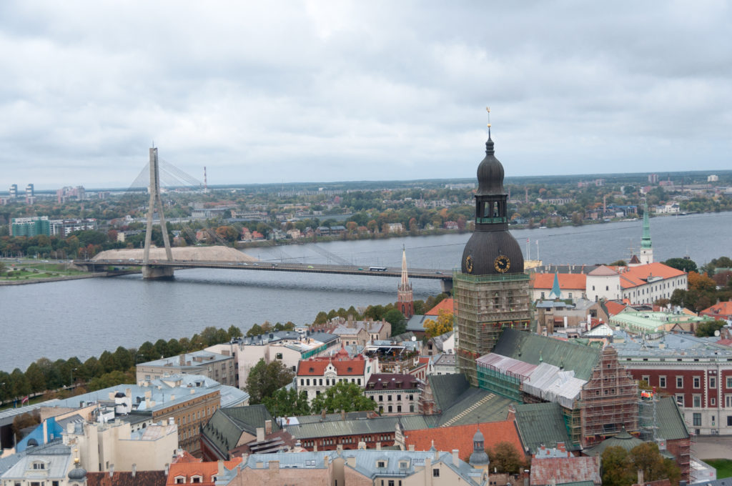 Old Riga rooftops and a dark domed tower with the Vanšu Bridge crossing the Daugava in the distance.