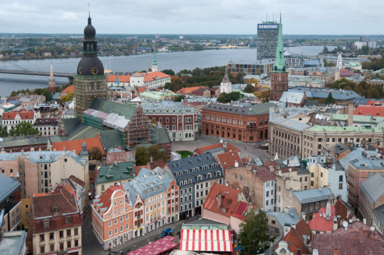 Elevated panorama of Old Riga rooftops and church towers with the Daugava River and modern city beyond.