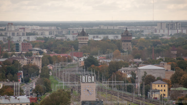 Elevated view over Riga railway tracks, a large RIGA clock sign, twin towers, autumn trees, and distant housing.