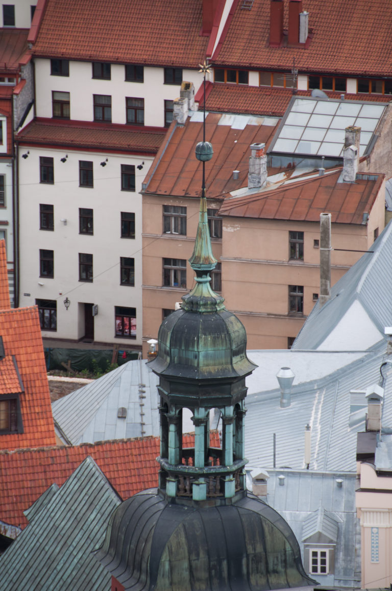 Green-patinated rooftop cupola and lantern rising among red-tile roofs, metal roofs, and chimneys in central Riga.