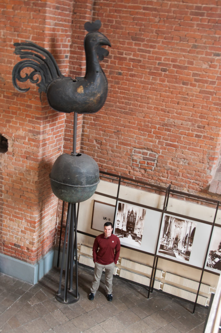 Large black metal rooster weather vane displayed indoors against brick, with a person and archival panels below.