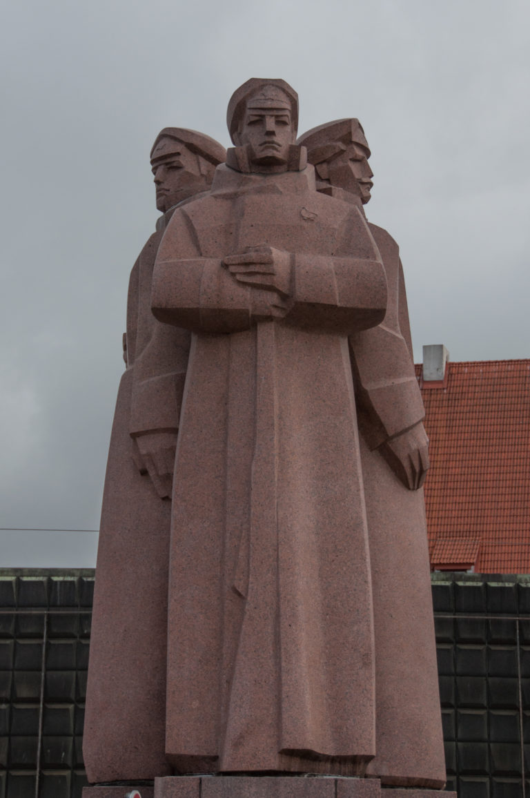Frontal close view of a red-granite group sculpture at the base of Riga’s Freedom Monument.