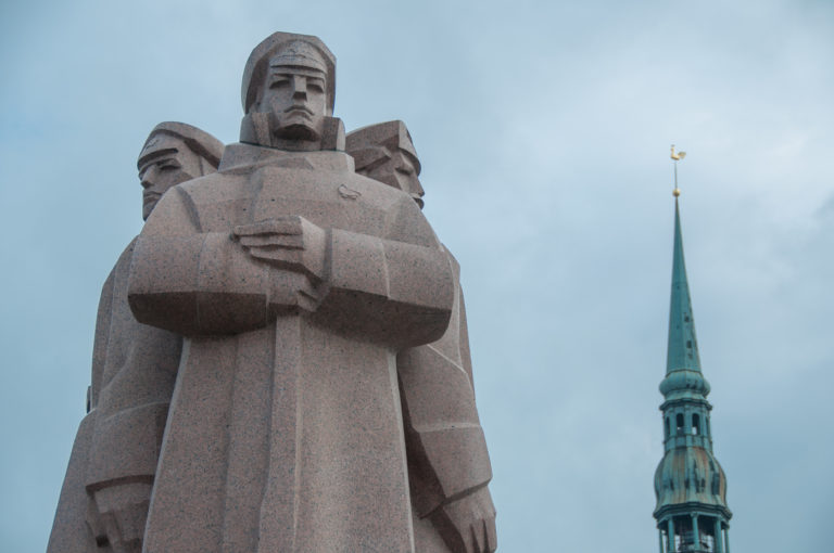 Low-angle view of a red-granite sculptural group at the Freedom Monument with a church spire behind.