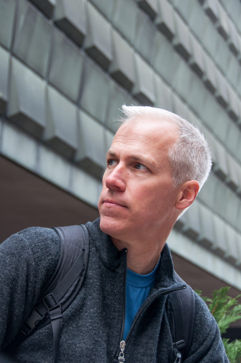 Close portrait of a man in a dark jacket against a faceted modern museum facade in Riga.