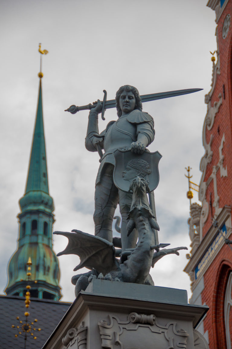 Rooftop sculpture of an armored warrior spearing a dragon, with Riga church spires and ornate facades behind.