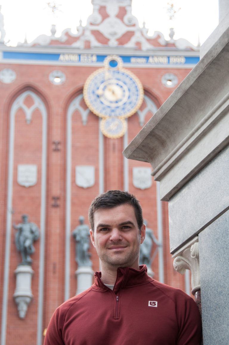 Man in burgundy top standing before the red-brick House of the Blackheads facade and clock in Riga.